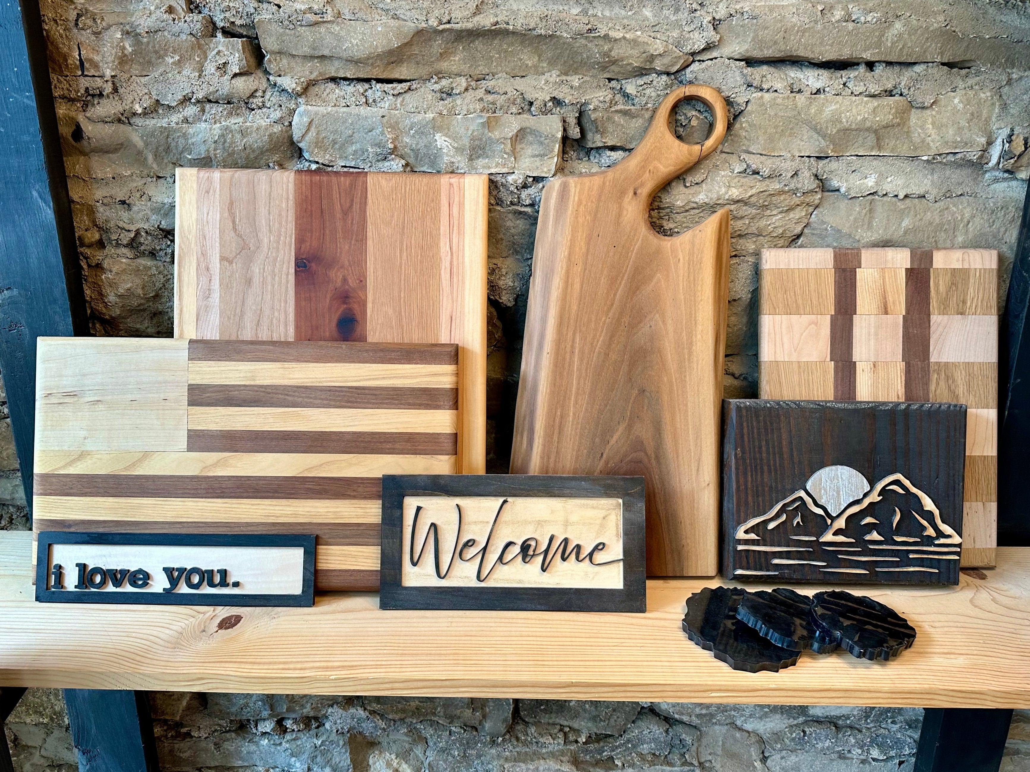 Collection of wooden cutting boards, charcuterie boards, coasters, and signs on a stone wall background