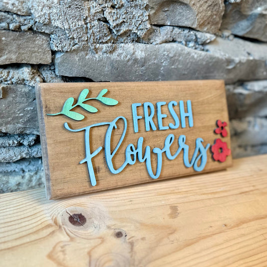 Wooden sign with 'Fresh Flowers' text and floral designs against a stone wall background