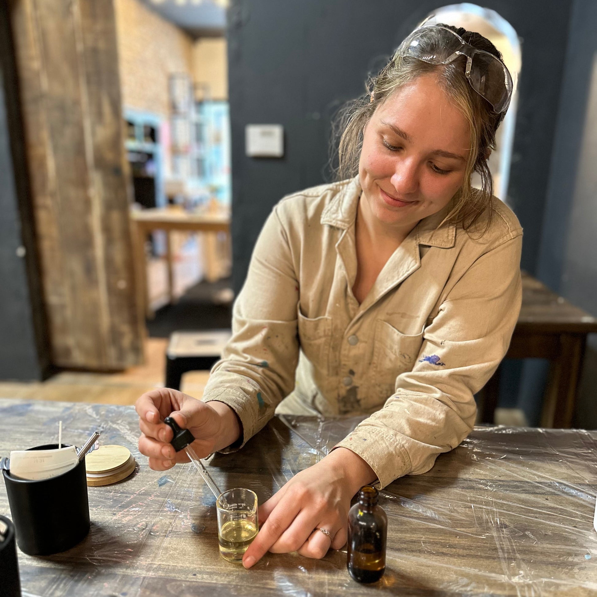 Person pouring essential oil from a dropper into a small glass container on a table with candle making supplies.
