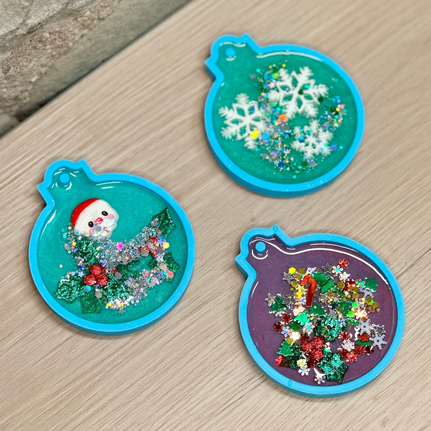 Three festive Christmas-themed epoxy ornaments on a wooden surface.