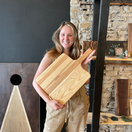 Woman holding a wooden pizza peel board in a room with stone walls and shelves.