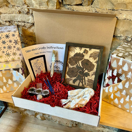 Box containing brushes, paint, instructions, and a framed artwork against a stone wall background
