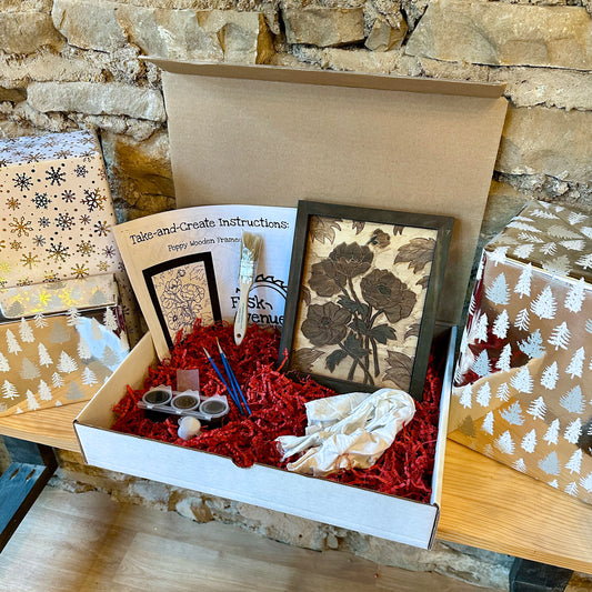 Box containing art supplies, instructions, and a wooden framed floral project against a stone wall background.