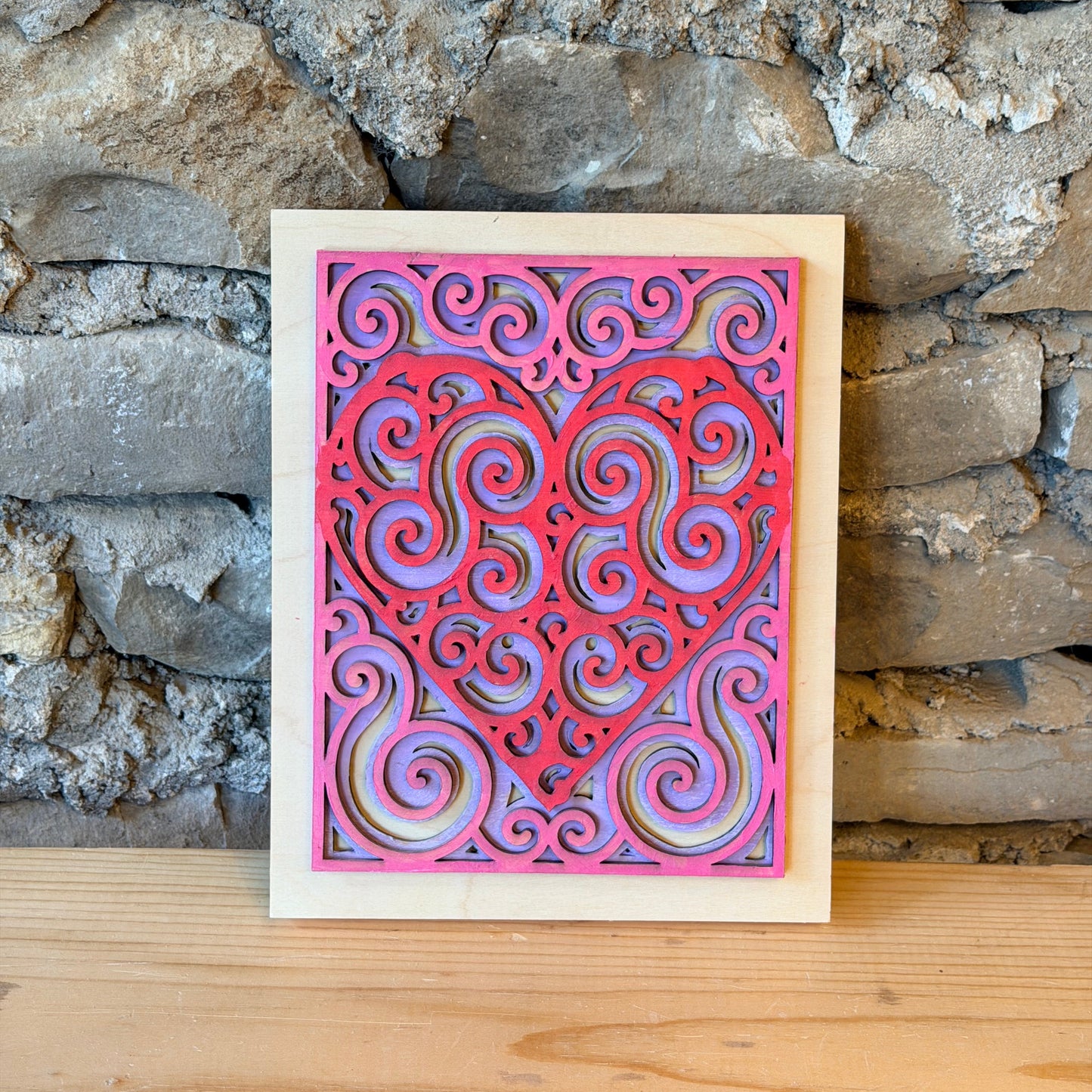 Decorative pink and purple heart mandala on a wooden backing against a stone wall.