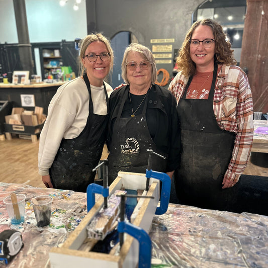 Three women in aprons standing behind a wooden workbench with an epoxy river coaster set in a mold on it.