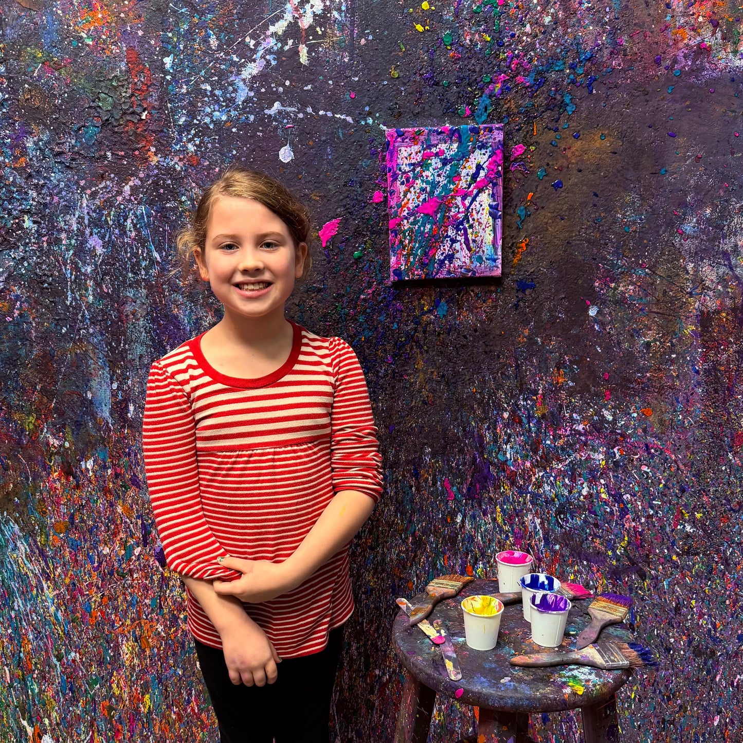 Young girl standing in an art studio with a colorful splatter painting on the wall.