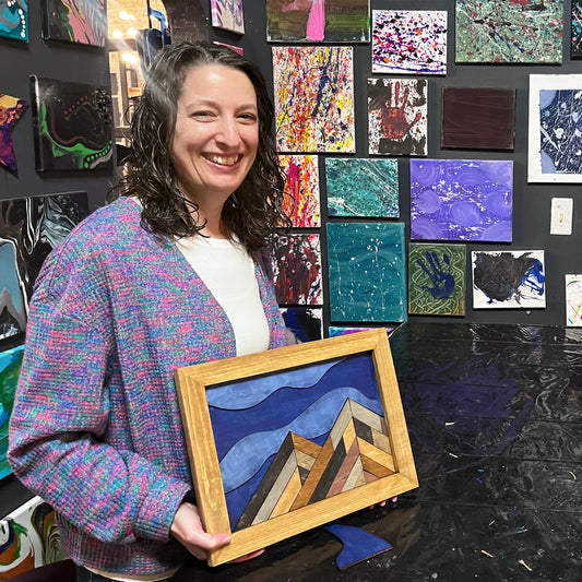 Woman standing in front of a wall of colorful art pieces, holding framed mountain artwork.