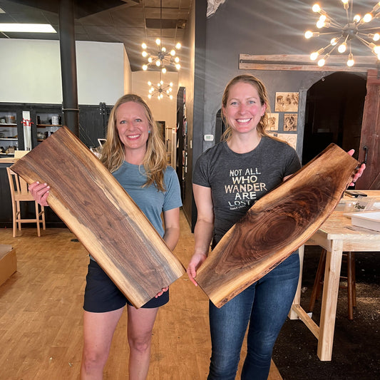 Two people holding lilve edge wooden trays in a room with wooden floors and a chandelier.