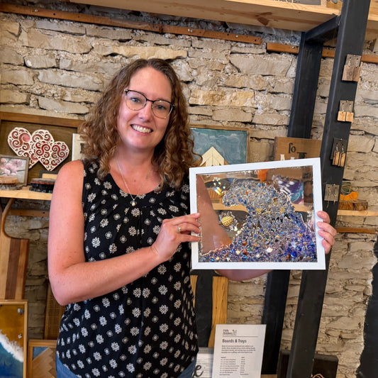 A woman in a studio holding up framed glass art. The artwork features a blue wave design with a turtle.