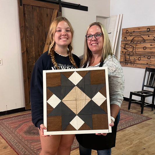 Two women holding a wooden checkered barn quilt board in a room with wooden flooring and furniture.