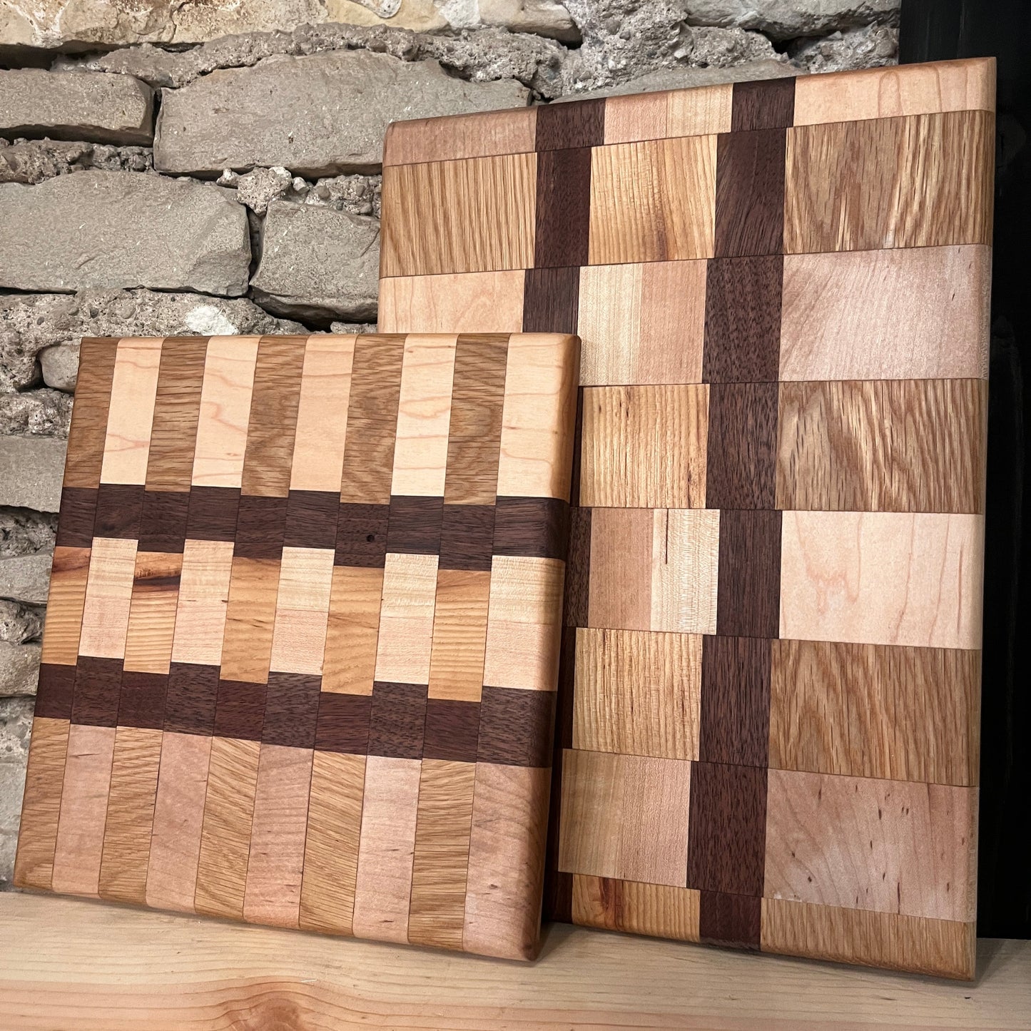 Two chaos-style cutting boards with a woven pattern of different wood colors placed on a wooden surface against a stone wall background.
