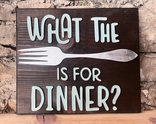 A wooden sign with the phrase 'WHAT THE FORK IS FOR DINNER?' painted in white, displayed against a brick wall.