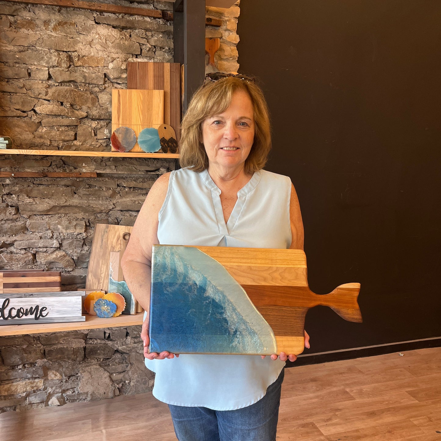 A woman standing in a storefront workshop setting, holding up a charcuterie board with a whale-shaped handle that is blue and has a wooden texture.