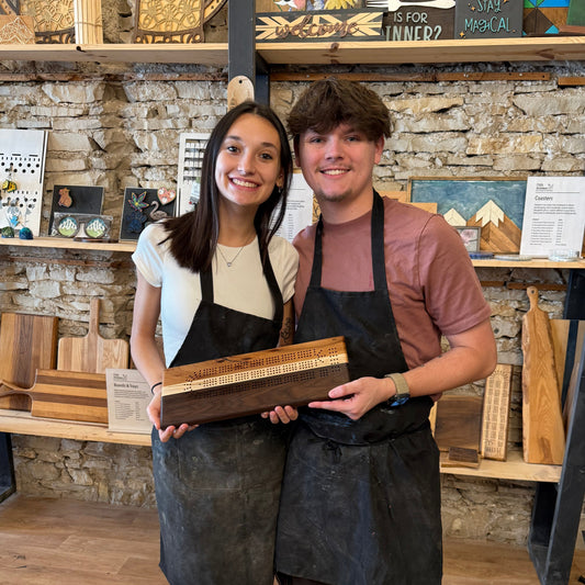 Two people standing in a rustic interior setting with a wooden cribbage board.
