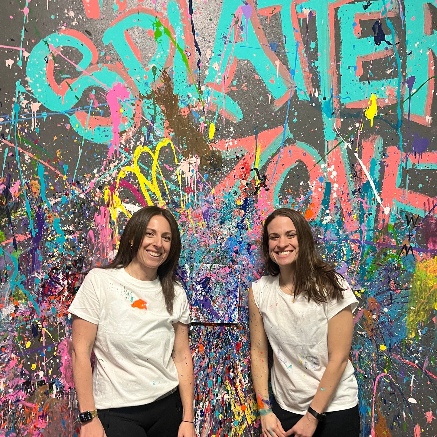 Two individuals standing in front of a colorful splatter paint artwork at a painting workshop.