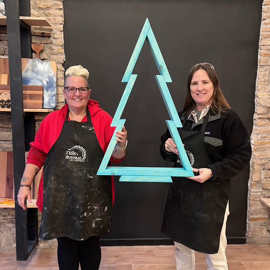 Two people holding a turquoise pine tree craft in a studio with stone walls and wooden floor.