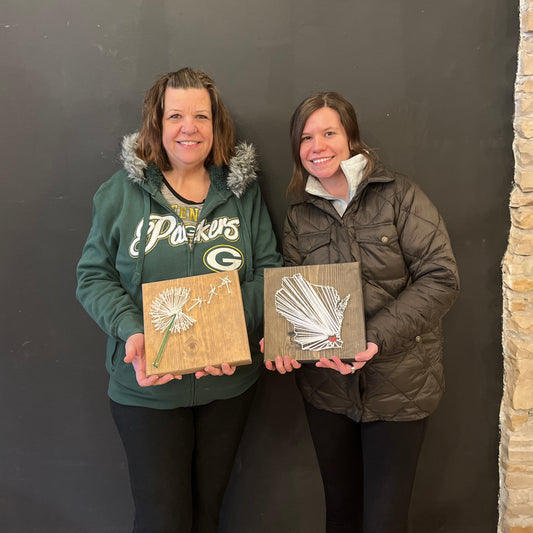 Two adults displaying their string art projects at a workshop. The projects are mounted on wooden boards with string designs in the center.
