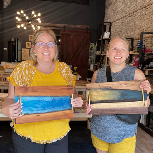 Two women holding wooden epoxy river serving trays with live edge wood in a workshop setting.