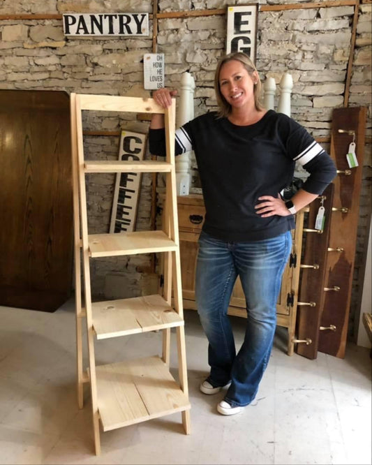 A person standing next to an unfinished wooden ladder shelf with multiple tiers, with a rustic appearance.
