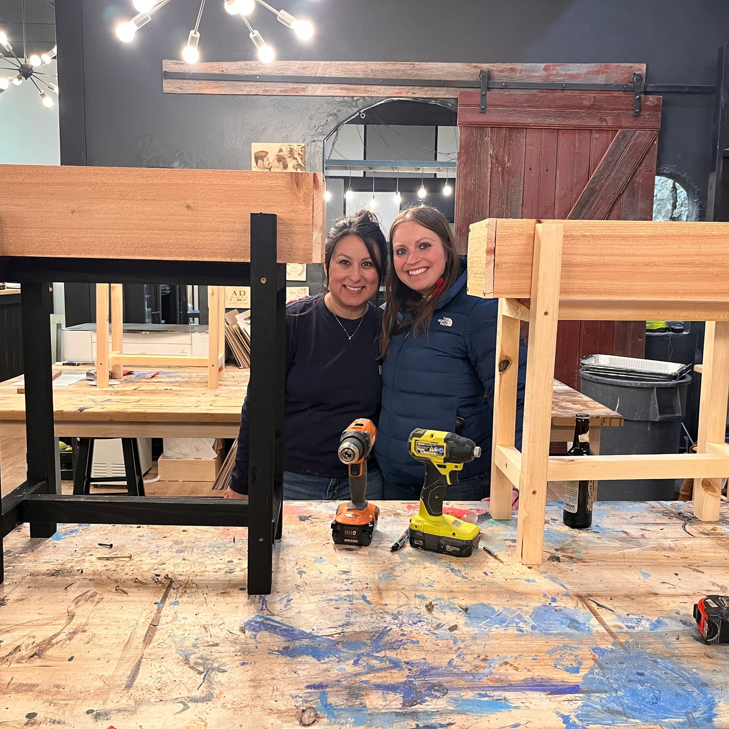 Two women posing with wooden planters in a workshop setting.