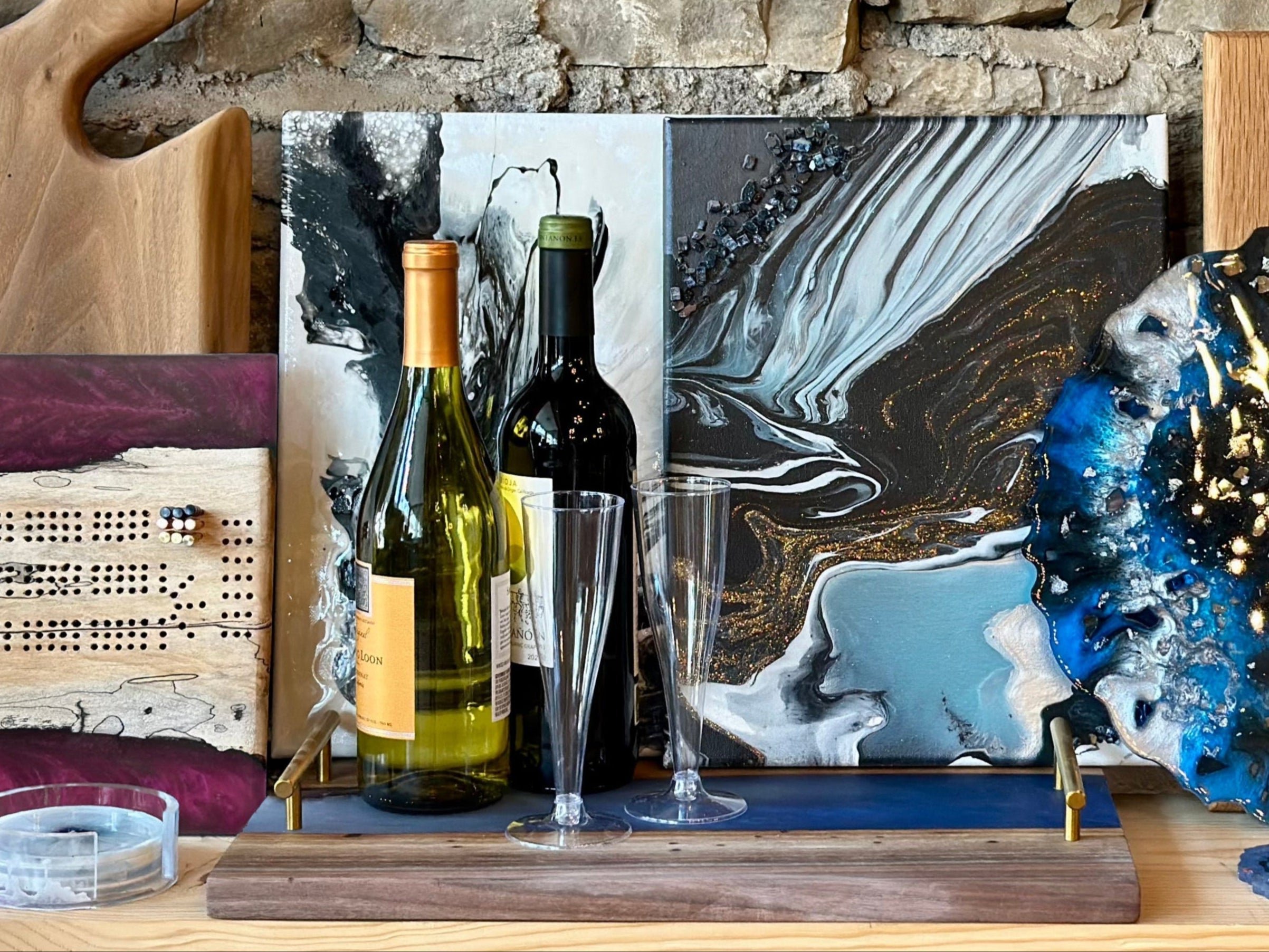 Two wine bottles with glasses on a wooden epoxy river serving tray in front of two abstract acrylic pour paintings, a wooden epoxy cribbage board, a charcuterie board, a cutting board, and an epoxy geode tray with epoxy coasters.