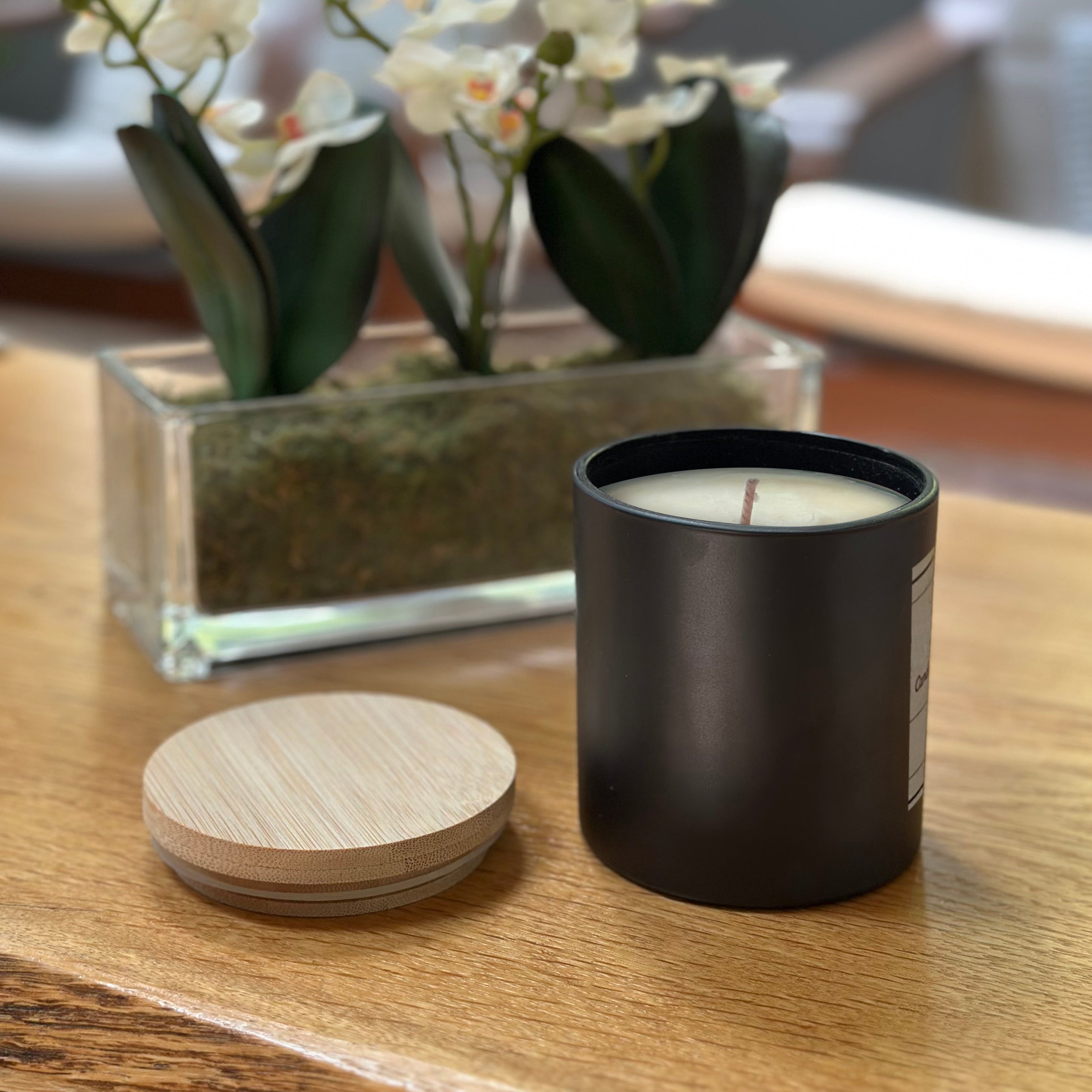 Black candle with wooden lid on a wooden surface, with a vase of flowers in the background.