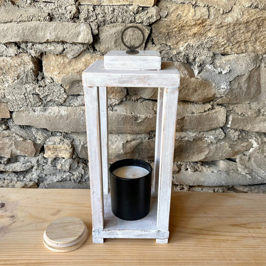White lantern with a black candle on a wooden shelf against a stone wall.