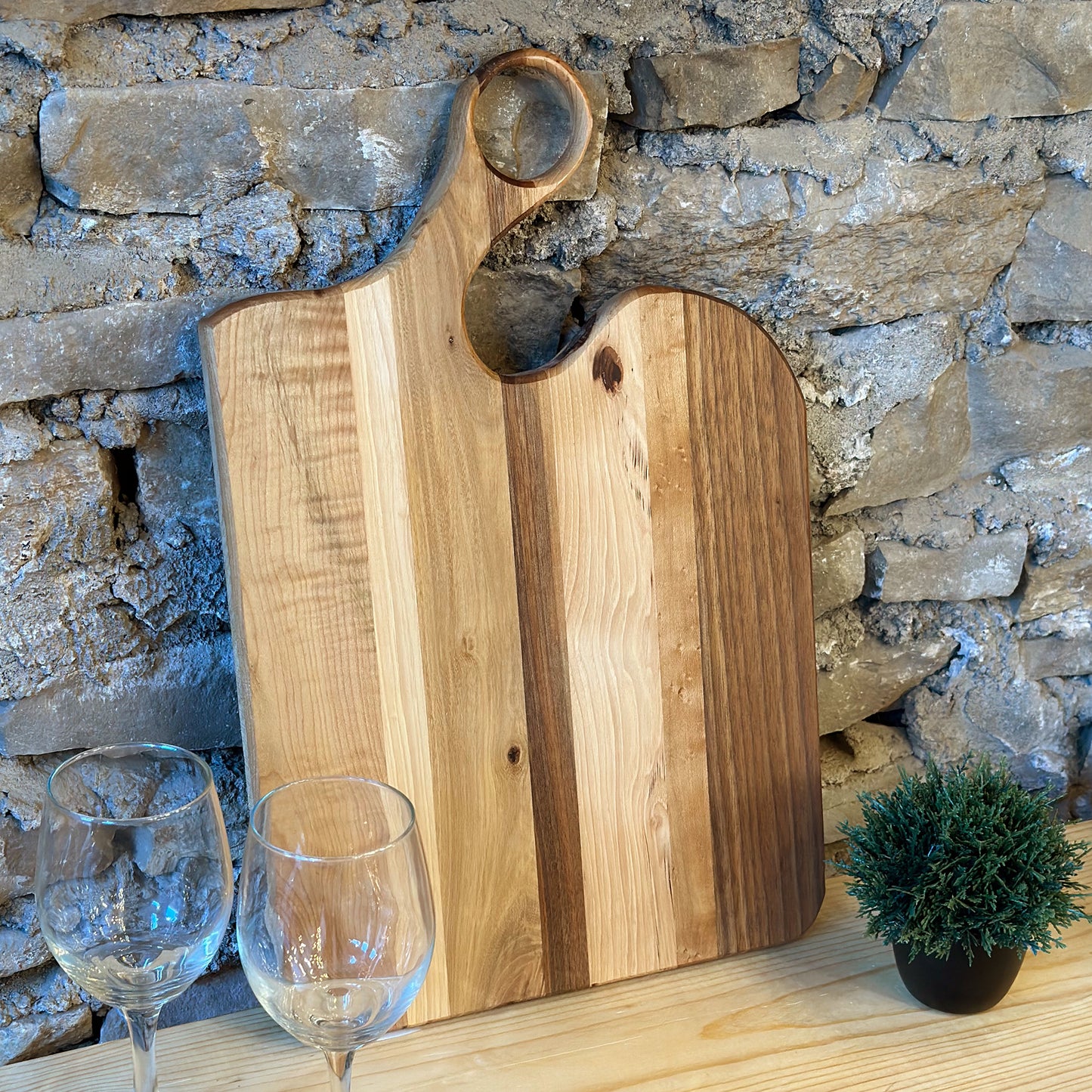 A walnut and maple wood charcuterie board with a striped handle, displayed on a wooden surface with a glass of wine and a small plant beside it.