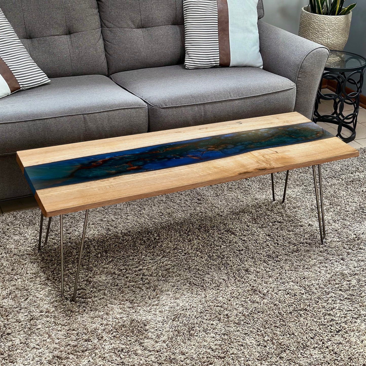 An epoxy river coffee table with live edge wood and metallic hairpin legs, featuring a blue and black epoxy pour, placed in a living room setting.