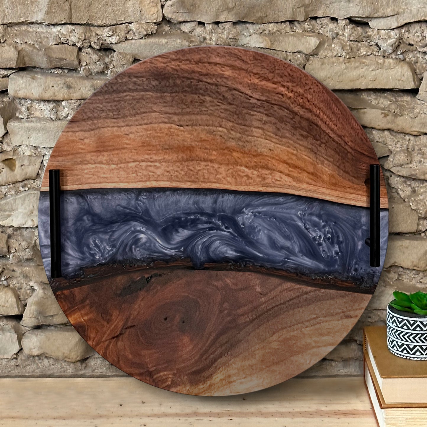 A round epoxy river tray with live edge wood on the edges and a swirling black and blue epoxy center, displayed on a stone surface.