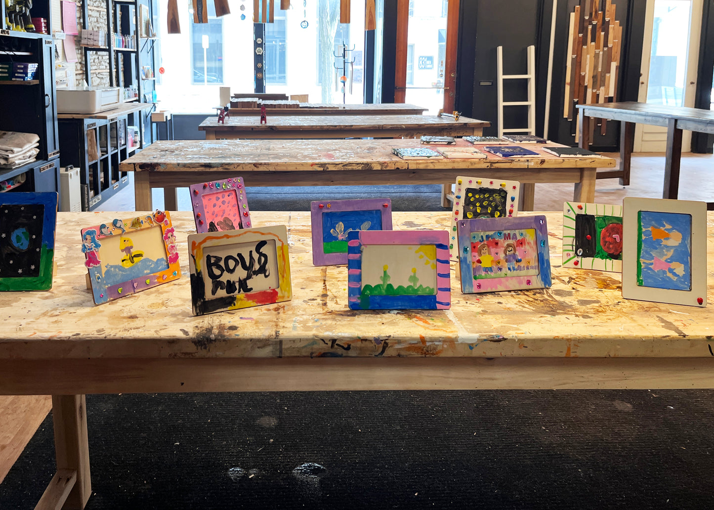 A series of homemade photo frames made by children, displayed on a table, with various decorative paints and tools visible in the background.