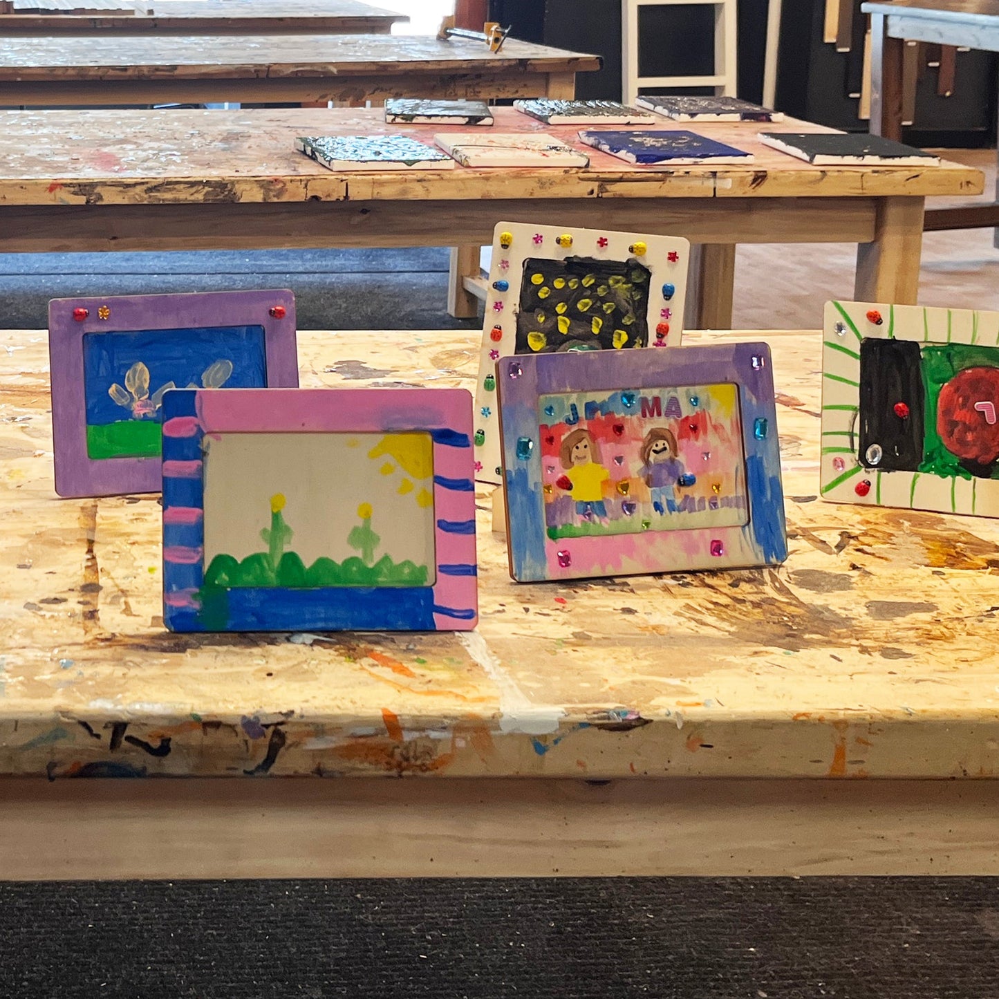 A series of homemade photo frames made by children, displayed on a table, with various decorative paints and tools visible in the background.