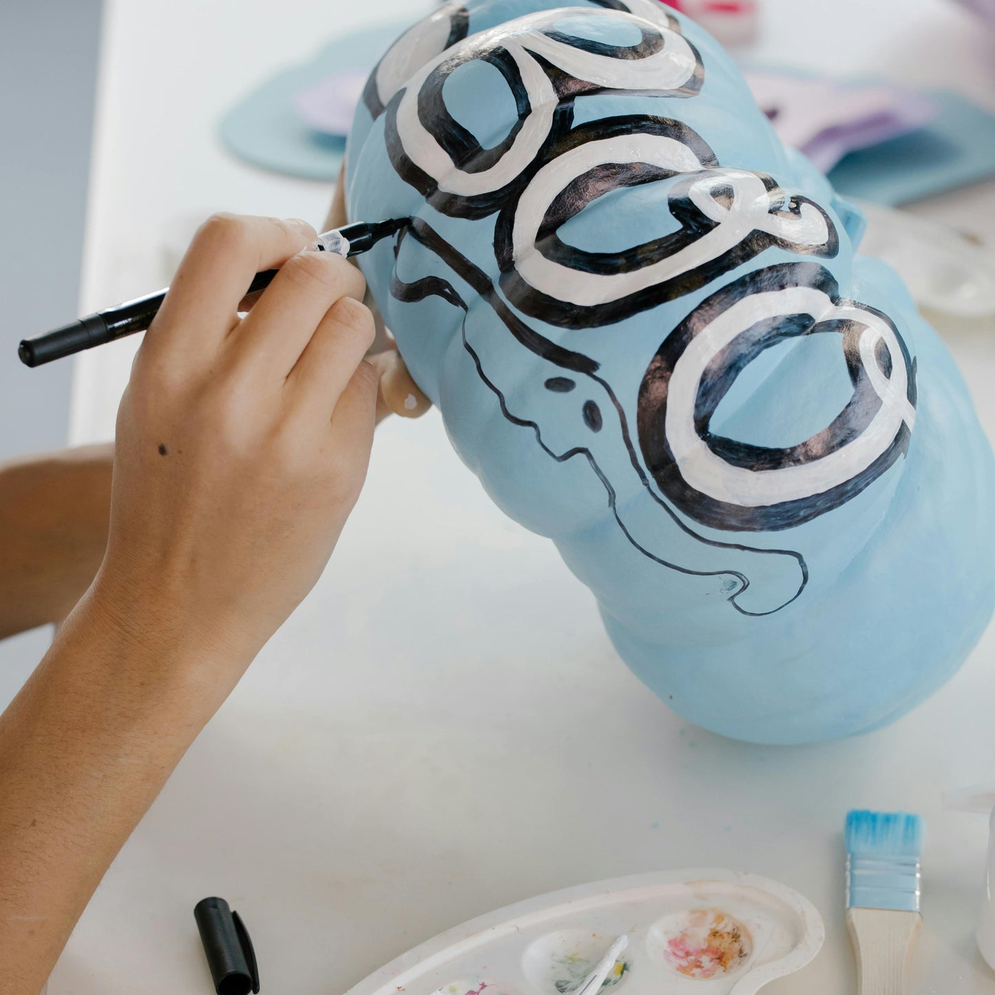 Person painting a blue pumpkin with the word 'BOO' and black ghosts designs on a white surface.
