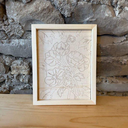 A rectangular natural wood frame containing a floral line art engraving, displayed on a wooden surface against a rustic stone wall background.