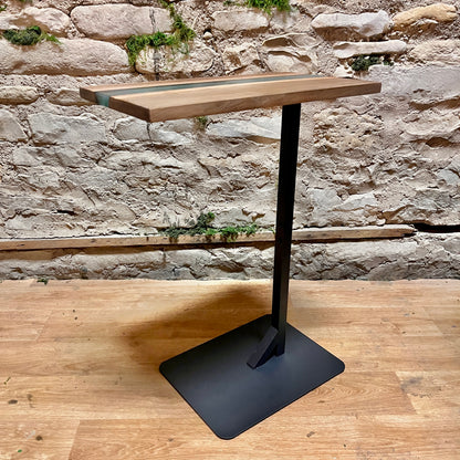 Wooden epoxy river side table with live edge wood and a black metal base against a stone wall.