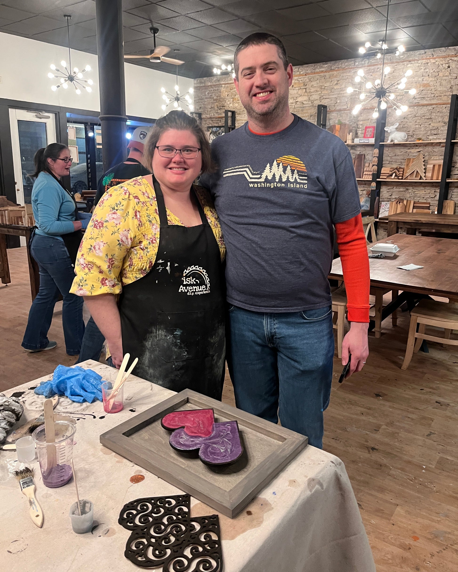 Two people standing in a studio setting with a table of epoxy heart wall decor craft materials