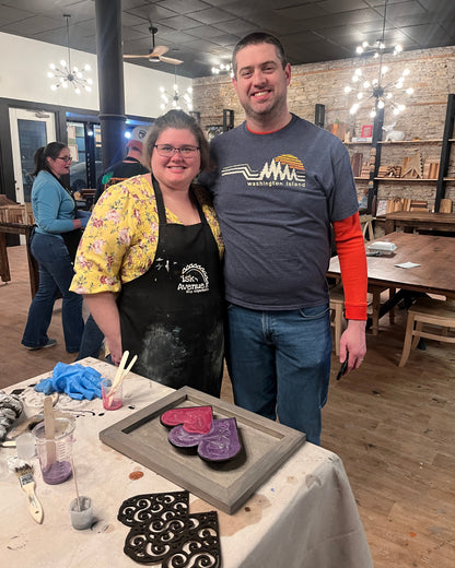 Two people standing in a studio setting with a table of epoxy heart wall decor craft materials