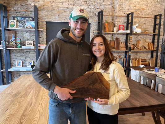 Two people holding a wooden live edge cribbage board in a studio room with shelves and decor.