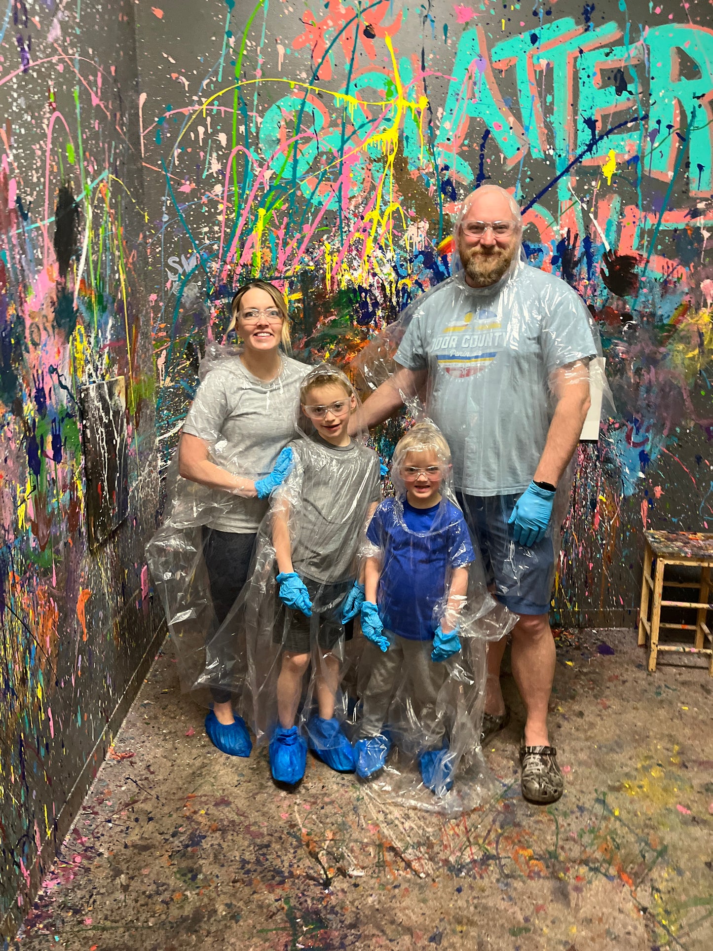 Two parents and two children wearing rain ponchos standing in a splatter room with colorful paint splatters.