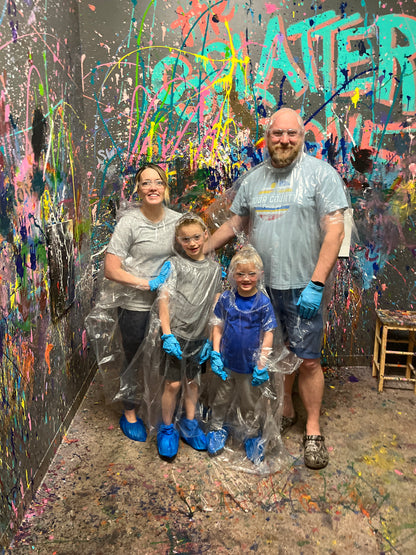 Two parents and two children wearing rain ponchos standing in a splatter room with colorful paint splatters.