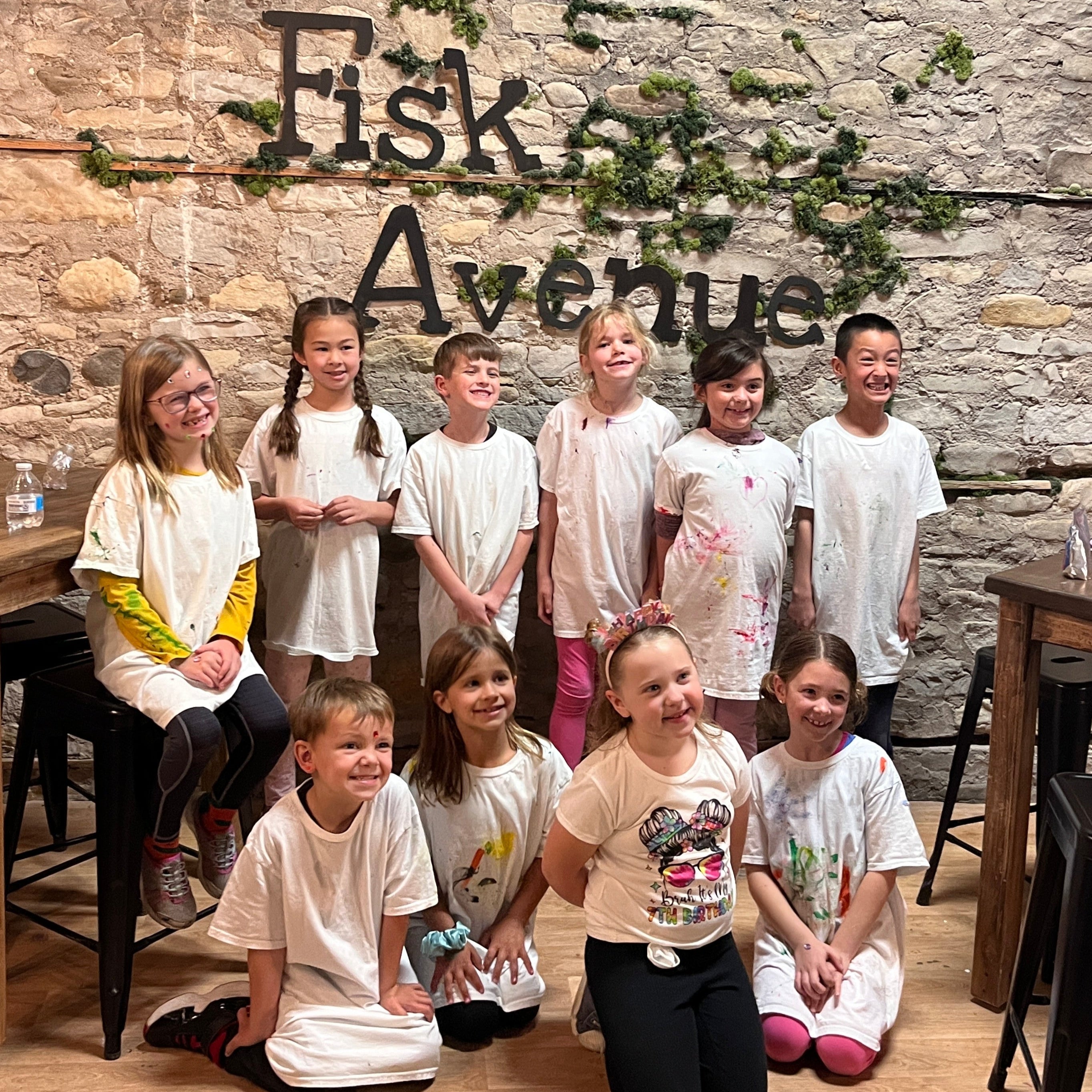 10 children wearing splatter painted shirts at a birthday party in a studio setting with 'Fisk Avenue' text and a stone wall