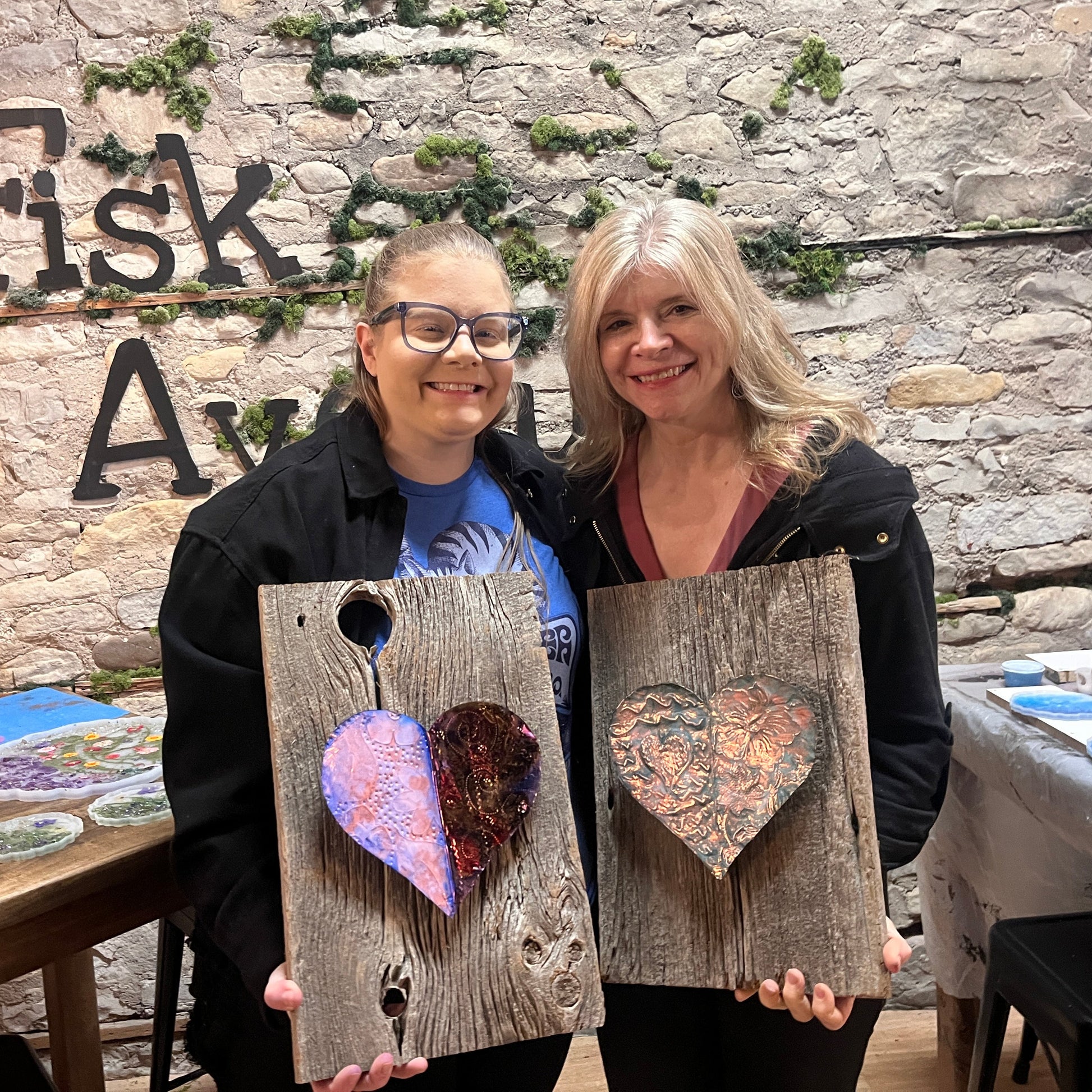 Two women holding rustic wooden boards with embossed copper heart designs in front of a stone wall with 'Fisk Avenue' text.