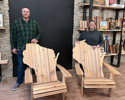 Two people standing with two wooden Adirondack outdoor chairs shaped like the state of Wisconsin in a store setting.