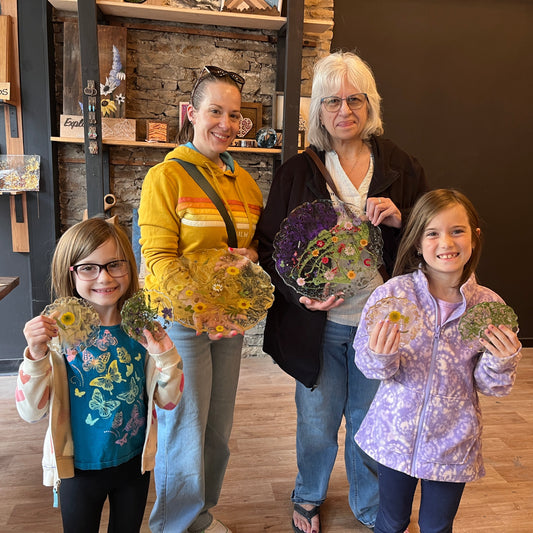 Four people, including two women and two girls, holding decorative floral epoxy serving trays and coasters with pressed flowers in a studio setting.