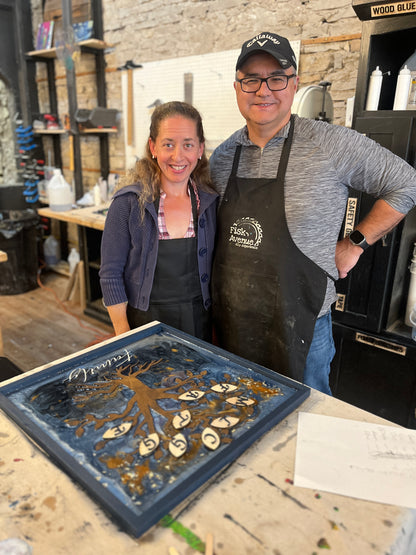 Two people in a workshop with a decorative wooden laser-cut family tree framed art piece with an epoxy background on a table