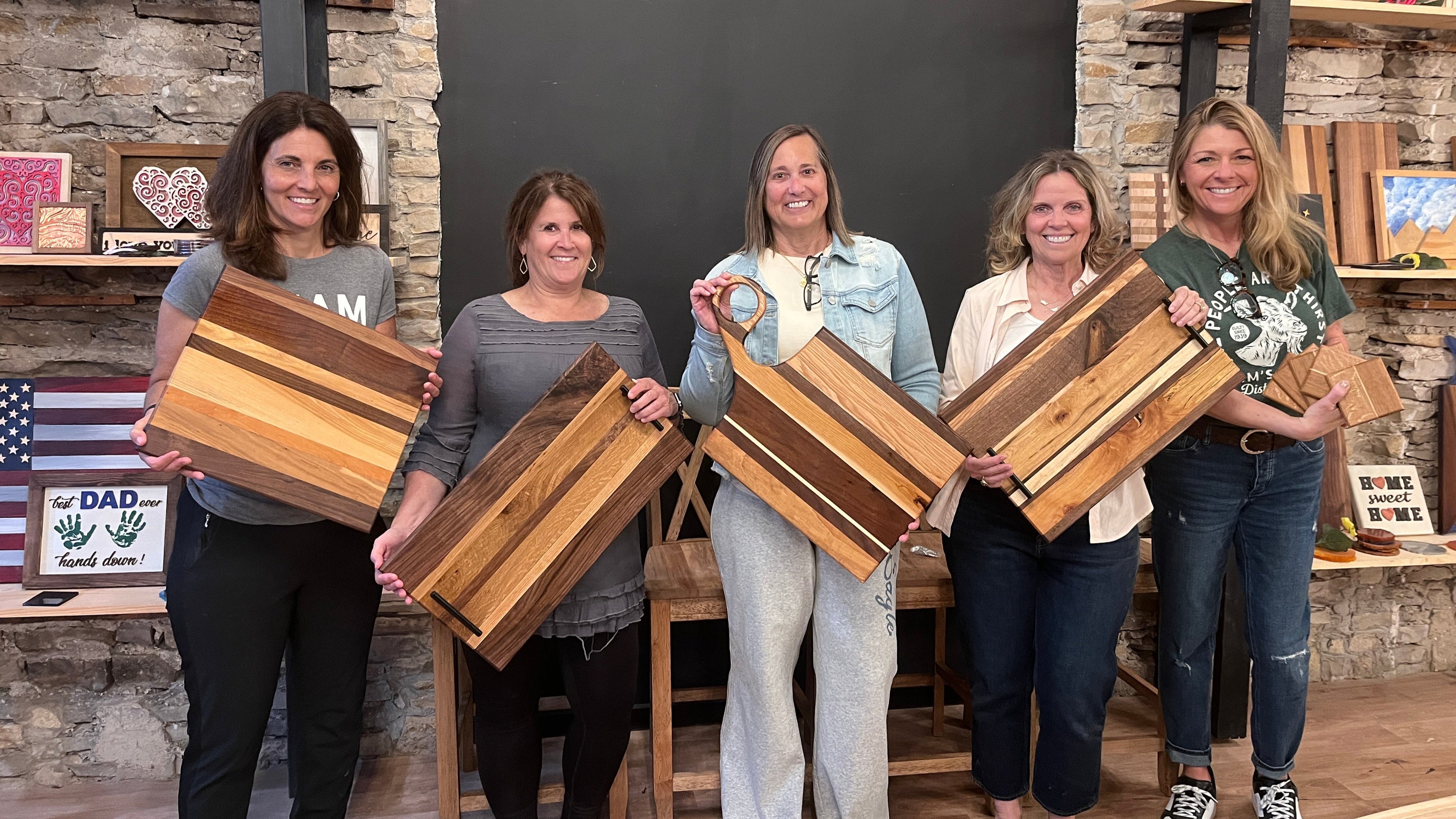 Five women holding wooden cutting boards and charcuterie boards in a studio setting with stone walls and crafts.