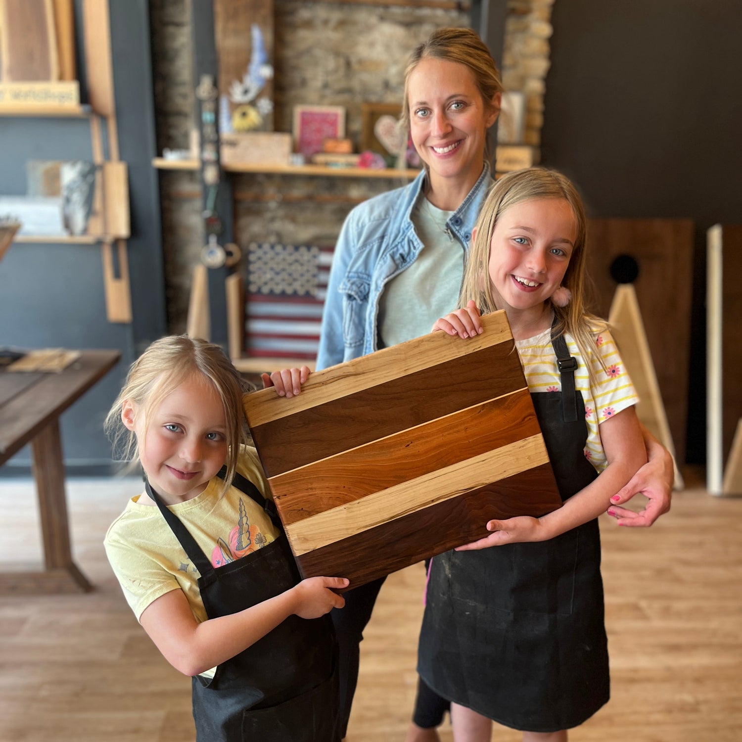 Two young girls and a woman holding a wooden cutting board in a workshop setting.