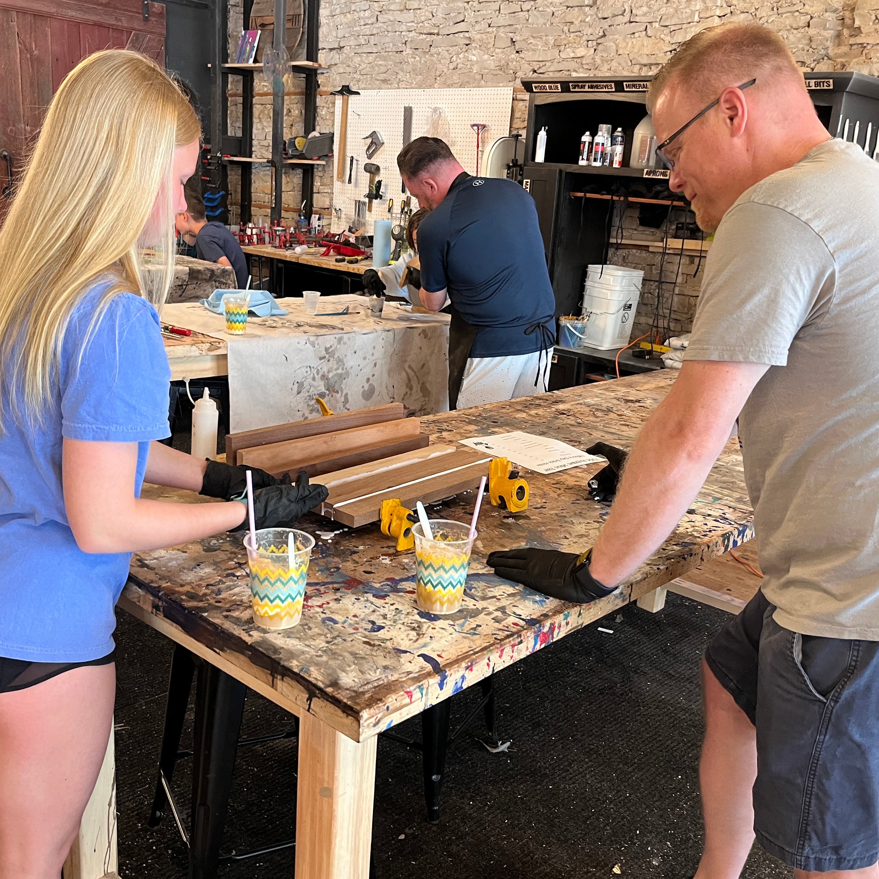 Two people working on a cutting board project at a table with various items in a workshop setting.