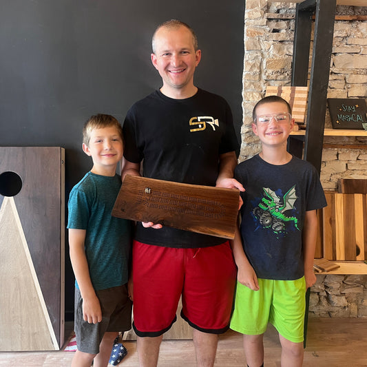 Man and two boys holding a wooden cribbage board in a studio with stone walls and shelves.