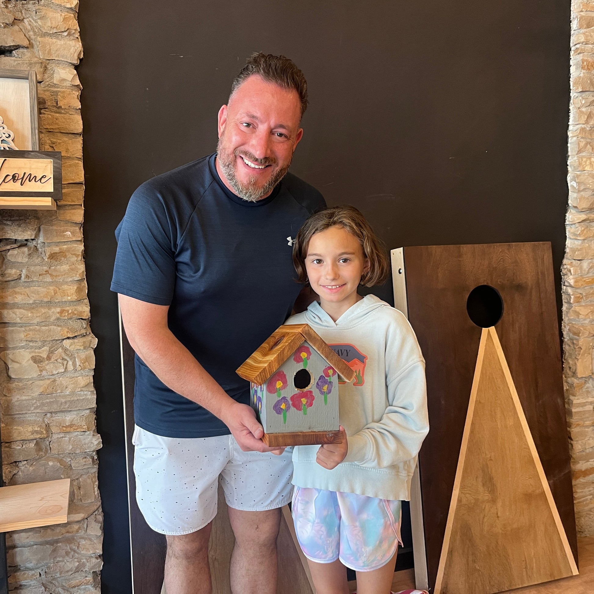 Man and young girl holding a wooden birdhouse with a decorative design, standing in front of a stone wall and wooden board.